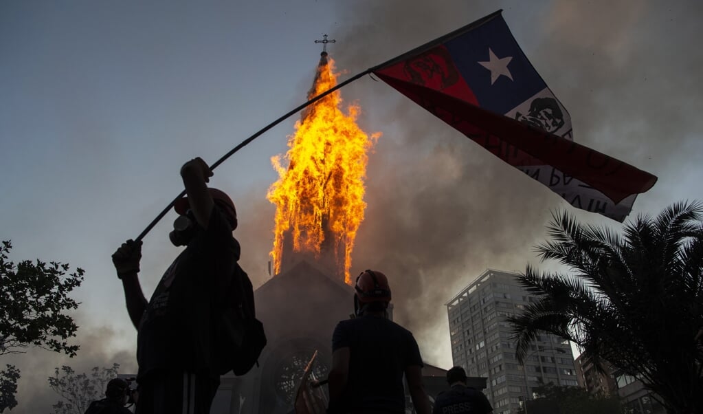 <p>Een actievoerder met de Chileense vlag bij de brandende toren van de kerk van Asuncion in Santiago, Chili. </p> (beeld afp / Martin Bernetti)