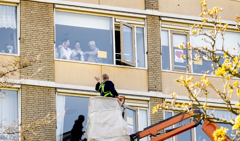 Een bezoeker zwaait vanuit een hoogwerker naar een familielid in het zorgcentrum. Gaan we van kwetsbaren echt vragen dat ze nog jaren geen bezoek mogen ontvangen? (beeld anp / Koen van Weel)