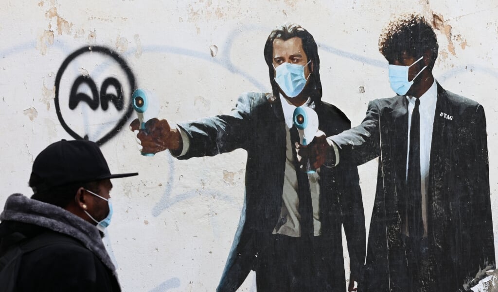 Een man in de Israëlische stad Tel Aviv loopt langs een muurschildering waarop de Amerikaanse acteurs John Travolta en Samuel L. Jackson staan. In plaats van wapens hebben ze thermometers in hun handen, die gebruikt worden om tijdens de coronapandemie op afstand te meten of mensen verhoging hebben. < (beeld afp / Emmanuel Dunand)