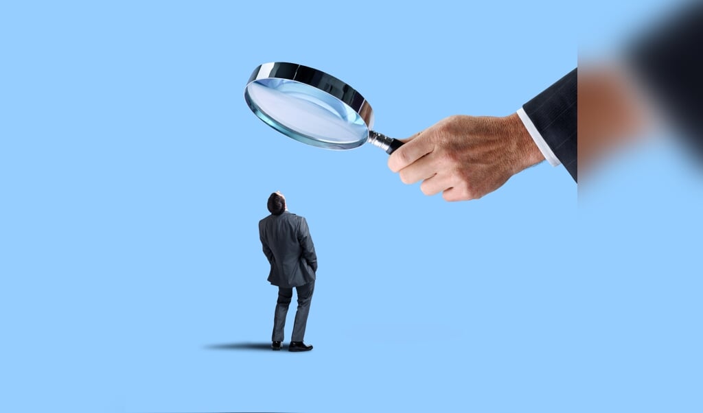 A businessman looks up at a large magnifying glass that is being held by an equally large hand isolated on a blue background. (beeld iStock)